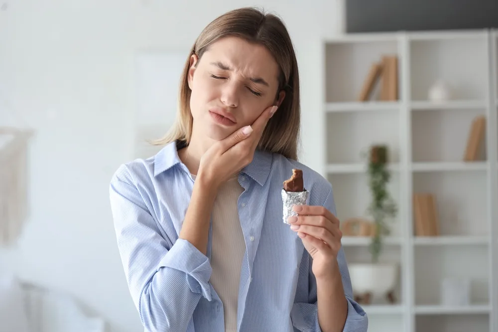 Woman holding a chocolate bar and touching her cheek in pain from tooth sensitivity